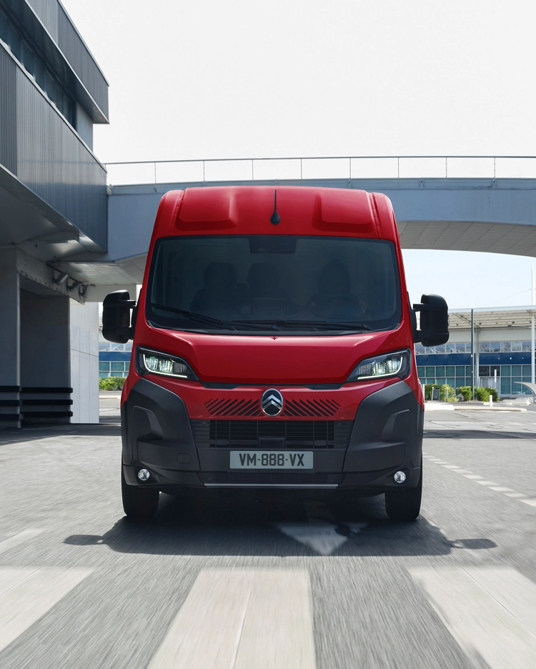 Vista frontal de una furgoneta Citroën Jumper roja circulando por un entorno urbano moderno, destacando su diseño robusto, practicidad y fuerte presencia en carretera.